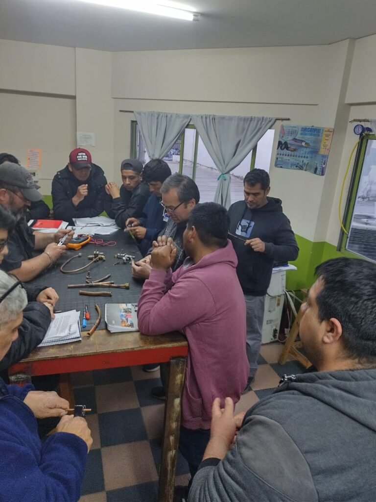 Grupo de estudiantes alrededor de una mesa de trabajo en el aula manipulando herramientas de pestaña, multímetros y tramos de caño de cobre durante una clase de climatización presencial