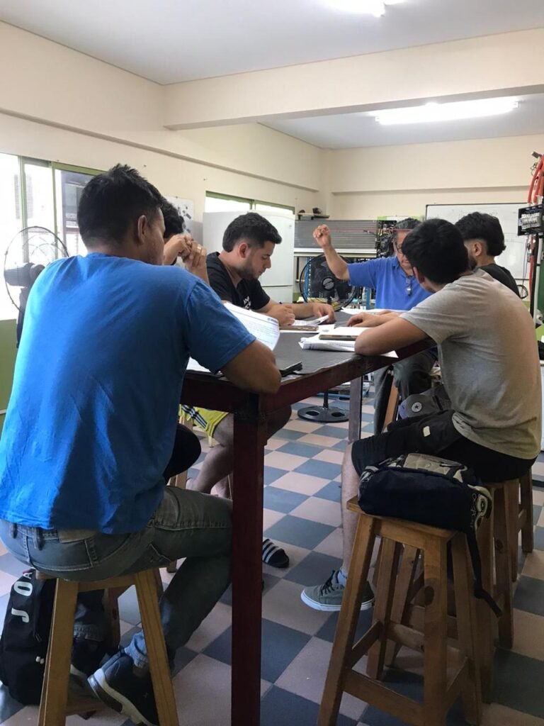 Grupo de alumnos sentados en banquetas de madera alrededor de la mesa de trabajo del aula técnica tomando apuntes mientras el profesor dicta los fundamentos de climatización