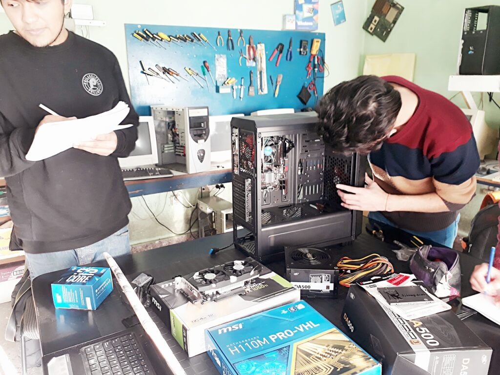 Estudiantes en el aula técnica instalando componentes de hardware como una placa madre MSI, fuente de alimentación y placa de video dentro de un gabinete de PC de escritorio