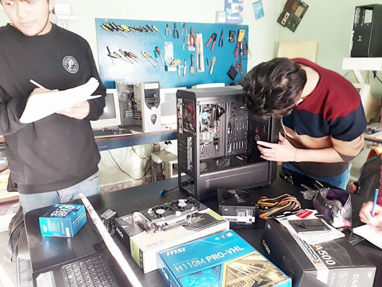 Estudiantes en el aula técnica instalando componentes de hardware como una placa madre MSI, fuente de alimentación y placa de video dentro de un gabinete de PC de escritorio