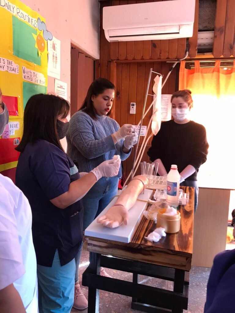Estudiantes y docente con guantes de látex y barbijo realizando una simulación de venopunción y preparación de jeringas sobre un brazo didáctico mecánico en el taller de Global-T