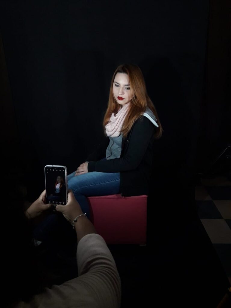 Estudiante tomando una fotografía con su celular a una modelo sentada en un puf rosa sobre un fondo negro en el estudio de fotografía