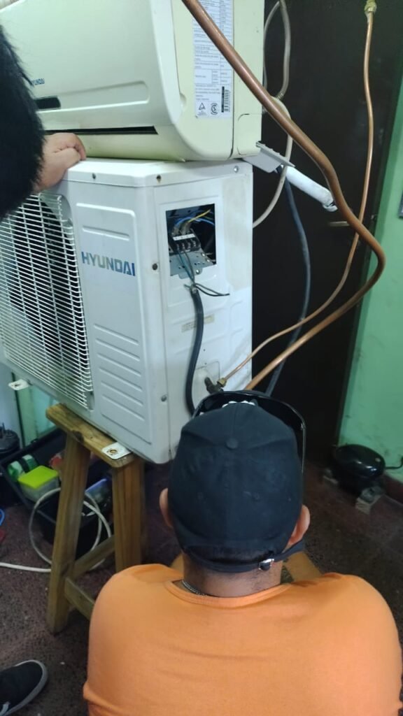 Estudiante de espalda con gorra negra trabajando en las conexiones eléctricas y cañerías de cobre de una unidad exterior condensadora split marca Hyundai en el taller didáctico