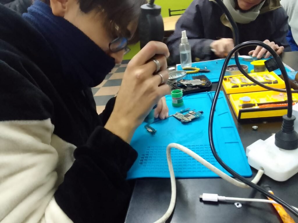 Estudiante utilizando una estación de aire caliente para desoldar componentes y pines de carga sobre una placa base de celular apoyada en una manta antiestática azul
