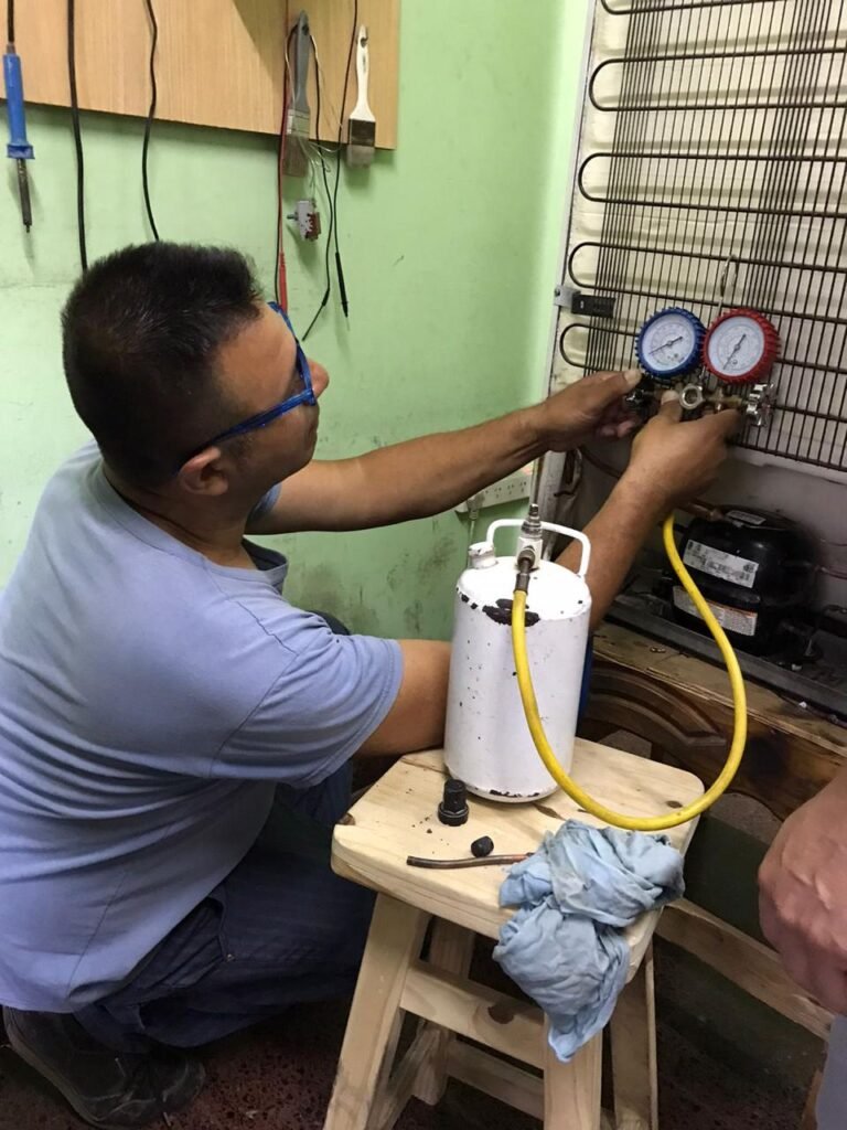 Estudiante con anteojos azules conectando un juego de manómetros (manifold) a la parte trasera de una heladera familiar junto a una garrafa de gas refrigerante blanca