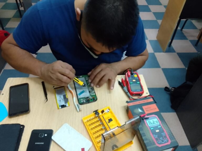 Estudiante utilizando un destornillador de precisión para desarmar un teléfono móvil expuesto junto a repuestos de baterías, pinzas de bruselas y un multímetro digital sobre el banco de trabajo