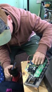 Alumno manipulando el conexionado de cables y relés en una placa electrónica pequeña mientras consulta el celular para verificar componentes