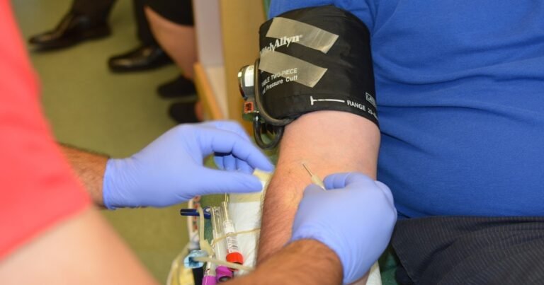 Primer plano de un enfermero utilizando guantes de látex azules para realizar una extracción de sangre con aguja en el brazo de un paciente que tiene colocado un brazalete de presión arterial WelchAllyn