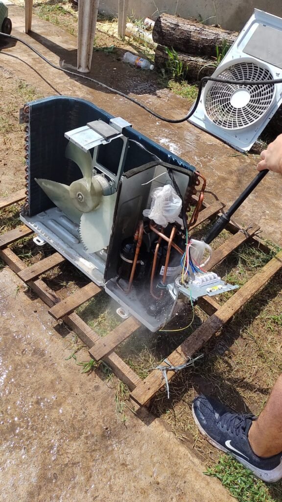 Técnico realizando una limpieza profunda con hidrolavadora a presión sobre las aletas del condensador de un equipo de aire acondicionado desarmado en el piso