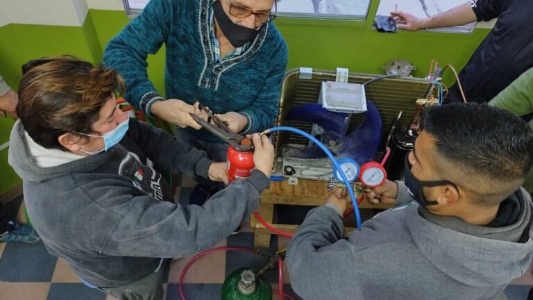 Vista aérea de alumnos y el instructor utilizando herramientas de corte, una garrafa verde de nitrógeno y un manómetro azul sobre una unidad condensadora desarmada en el aula