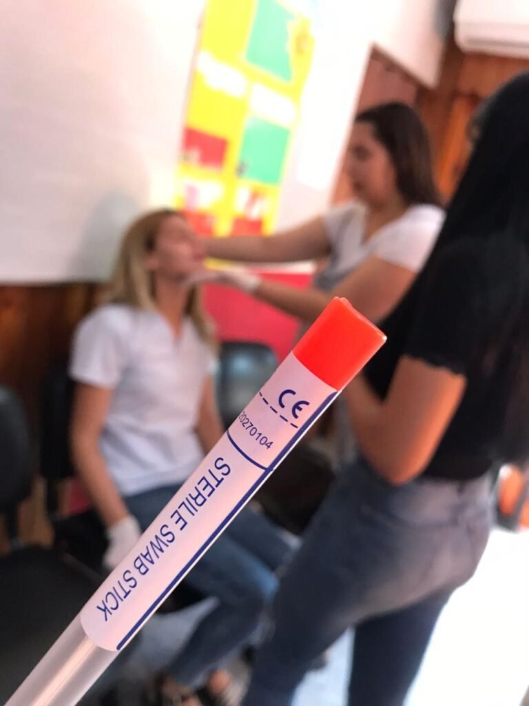 Tubo con etiqueta Sterile Swab Stick en primer plano enfocado mientras al fondo, de forma difuminada, dos estudiantes realizan una simulación de hisopado en el aula técnica