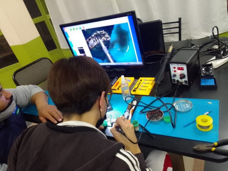 Alumno de espalda trabajando con soldador tipo lápiz frente a un monitor LED que muestra la imagen ampliada de un circuito integrado por microscopio digital junto a una estación Kushiro