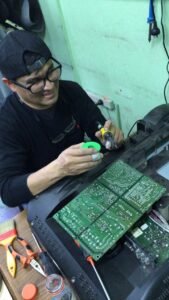 Estudiante con anteojos sonriendo mientras sostiene un soldador tipo lápiz y estaño para reparar las pistas y soldaduras de una placa de circuito integrada grande