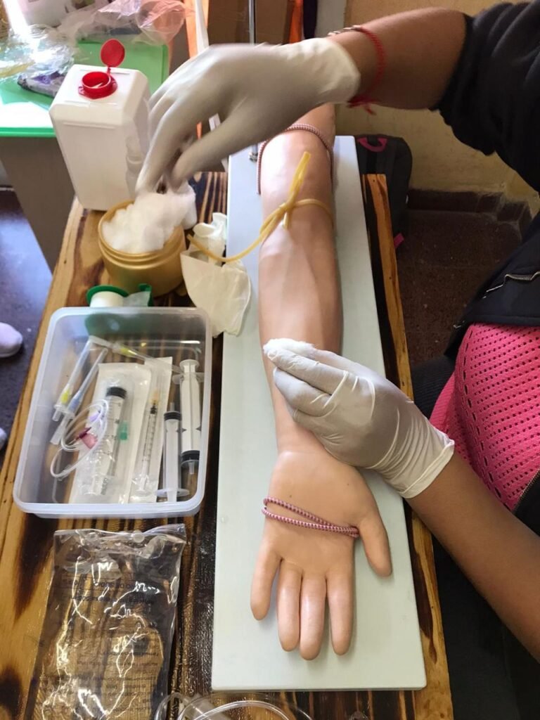 Primer plano de manos con guantes blancos utilizando algodón para limpiar la zona de punción en un brazo simulador con lazo descartable, junto a una bandeja con jeringas, agujas y tubos de ensayo,